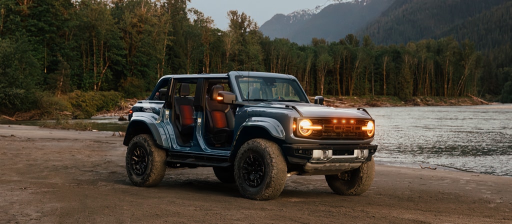 Un VUS Ford Bronco® 2025 avec les portes retirées et les lumières allumées, stationné près d’un lac dans la forêt
