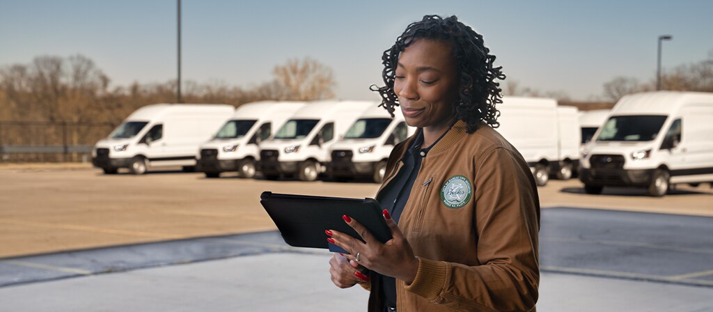Un parc automobile de véhicules Ford E-Transit™ 2025 blancs stationnés derrière un travailleur tenant une tablette