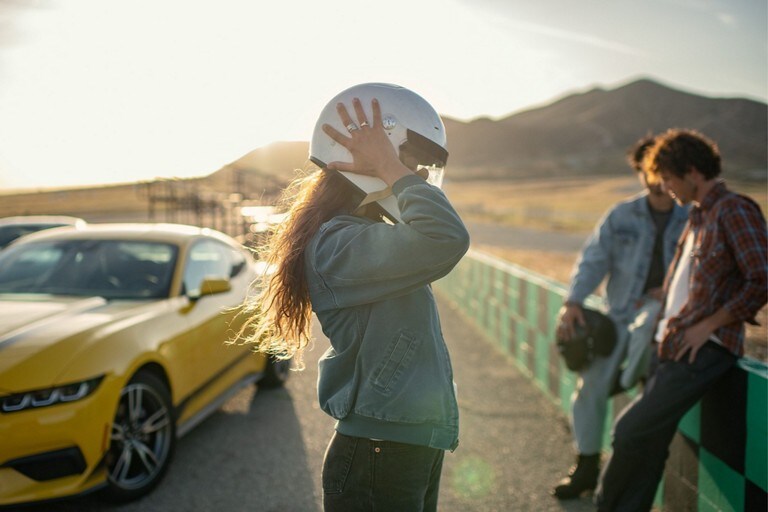 Une femme enfile un casque de course sur une piste. Deux hommes et une Ford Mustang sont en arrière-plan. 