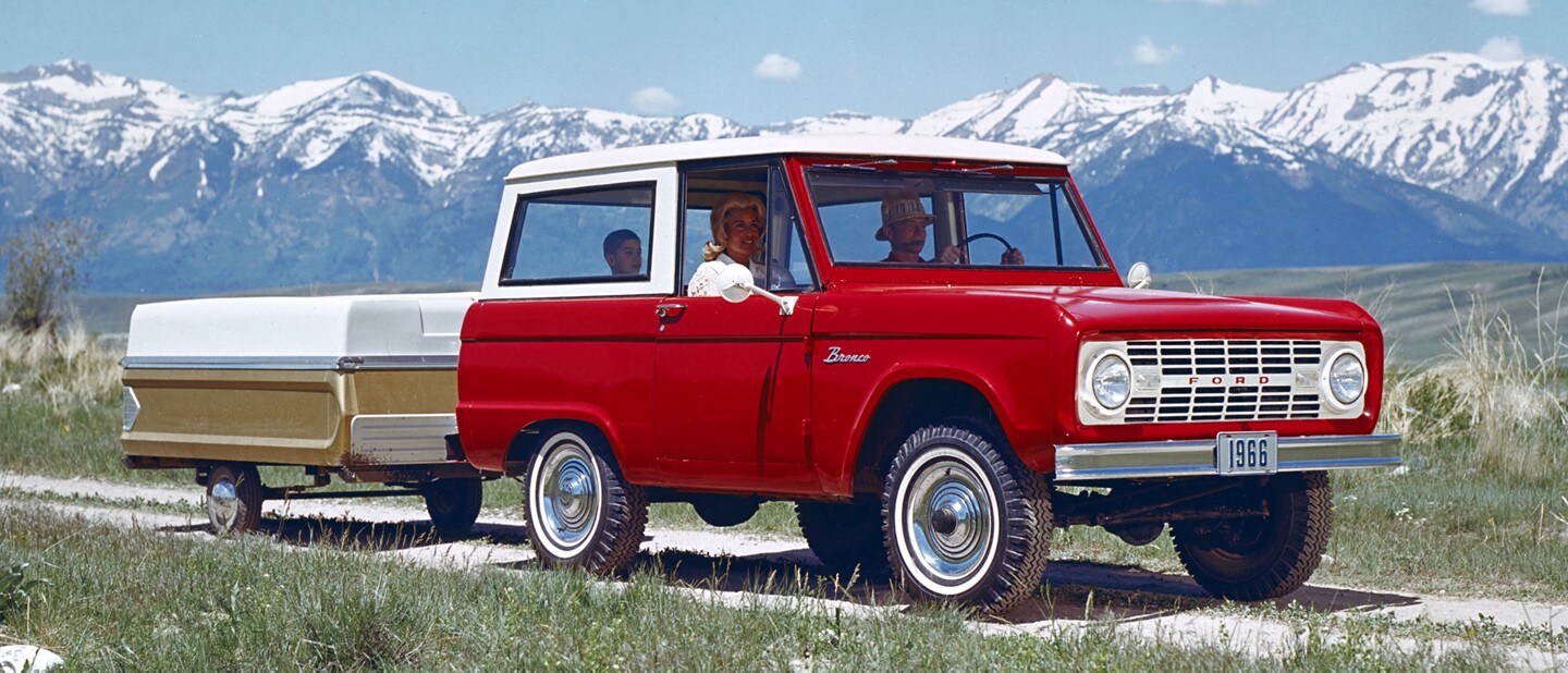 Une famille en route dans un fourgon Ford Bronco Sport 1966 en Royal Maroon avec un toit blanc Wimbledon tirant une remorque de camping dans les montagnes