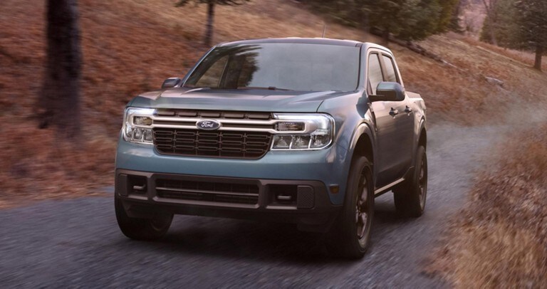 Ford® Maverick LARIAT 2022 de couleur Zone 51 avec l'ensemble tout-terrain FX4 en option circulant sur une route bordée de conifères au crépuscule