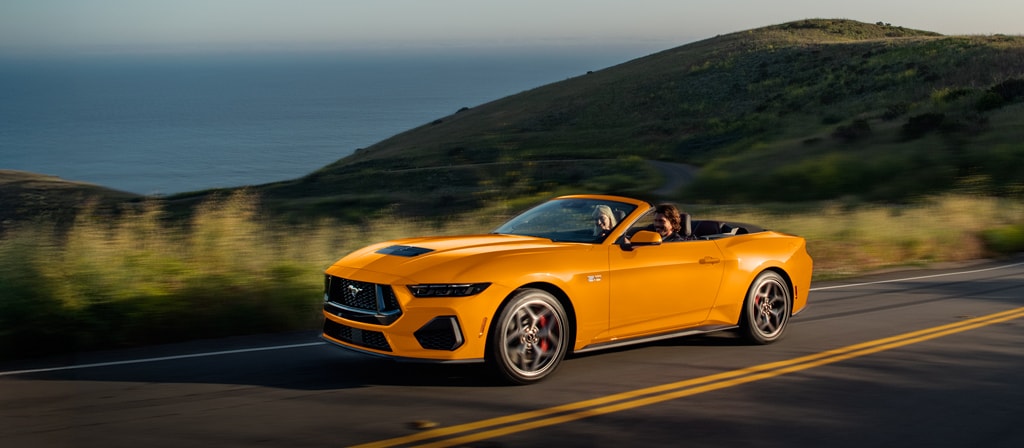 Deux personnes dans une Ford Mustang® GT décapotable 2026 couleur Furie orangée métallisée à trois couches circulant sur une autoroute côtière