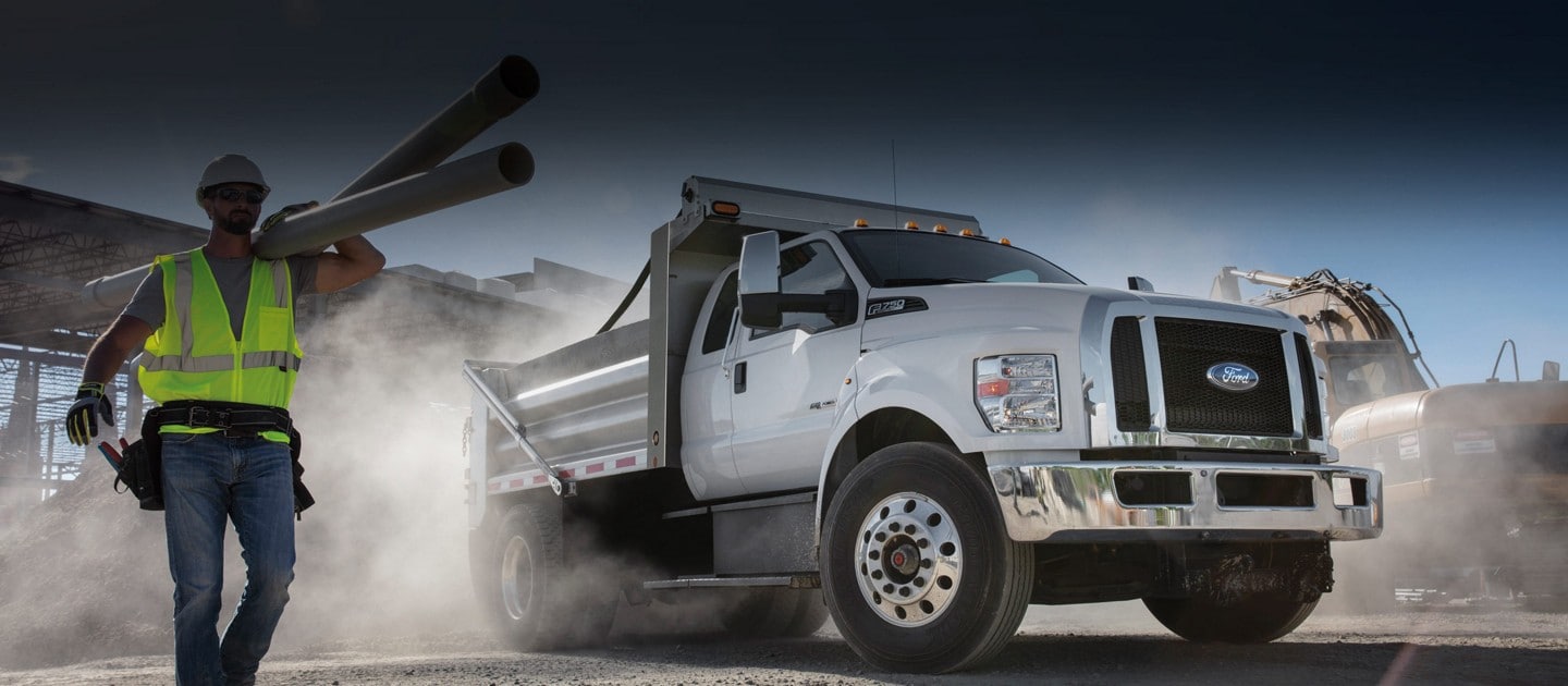 Un Ford F-750 2025 avec équipement de camion-benne en blanc Oxford conduit près d'un ouvrier sur un chantier de construction poussiéreux