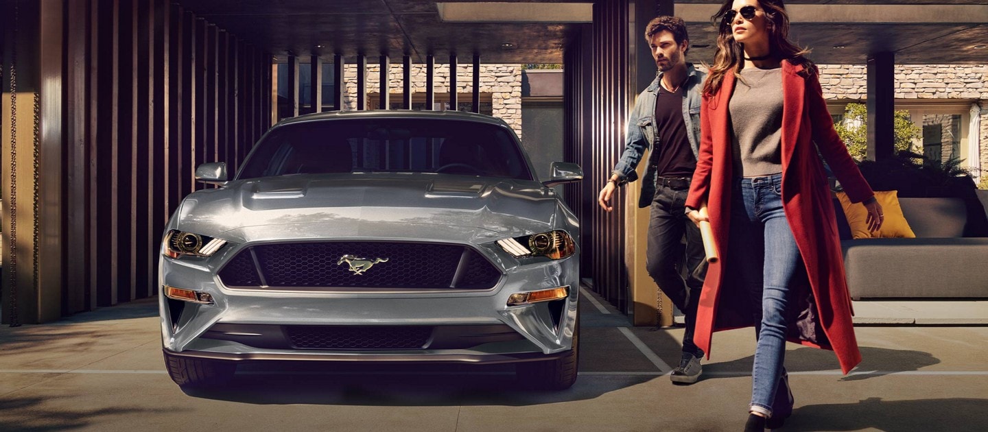 Coupé Ford Mustang® 2023 en gris carbonisé métallisé stationné dans un port d’auto avec deux personnes s’éloignant
