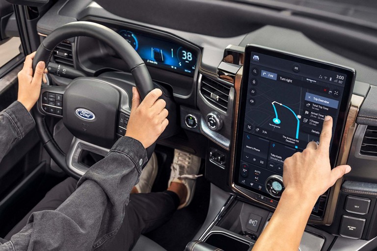 Une personne conduit un véhicule Ford tandis que le passager interagit avec l’écran de navigation connectée.