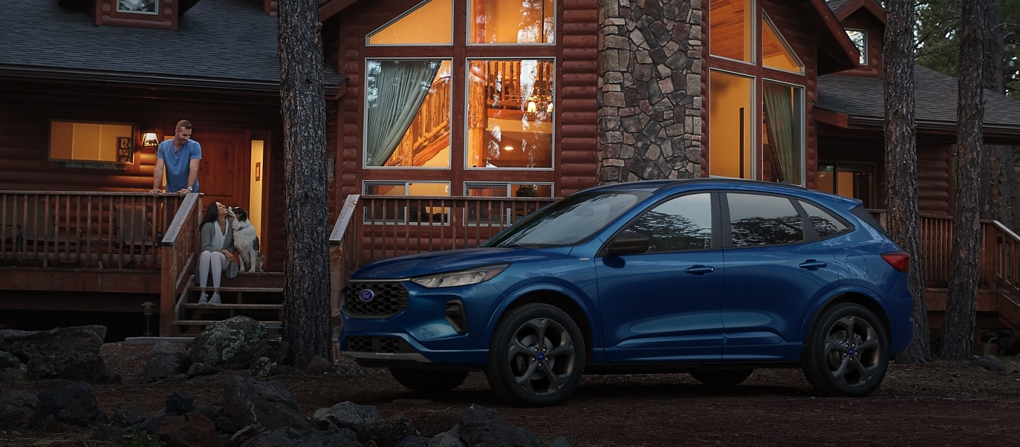 Un VUS Ford Escape bleu stationné devant une grande maison dans une forêt. Un couple et son chien sont assis sur une galerie.