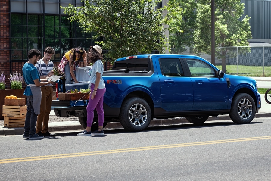 Un groupe de personnes charge des plantes sur le plateau d’un camion Ford Maverick bleu stationné en bordure de trottoir.