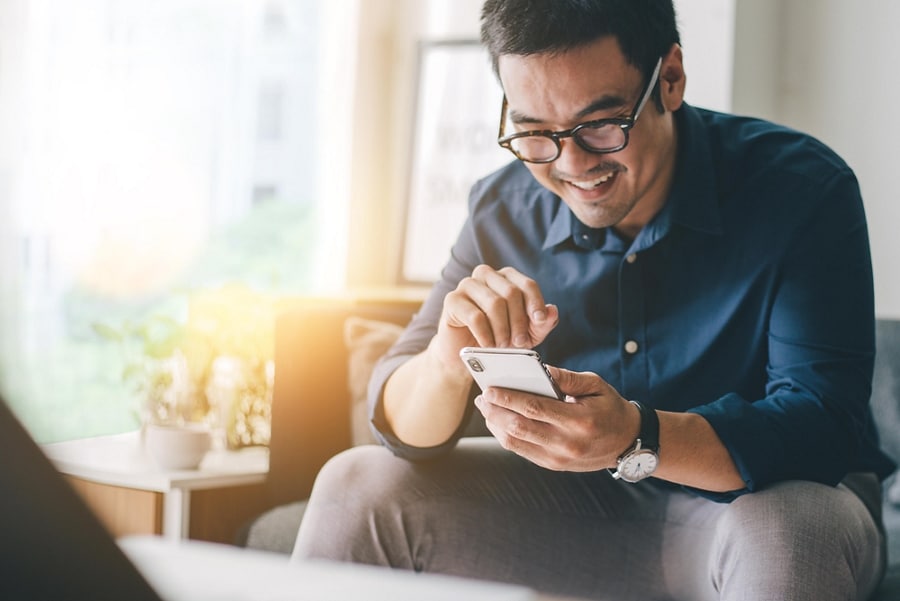 Un homme assis dans un salon ensoleillé sourit en regardant son téléphone intelligent.