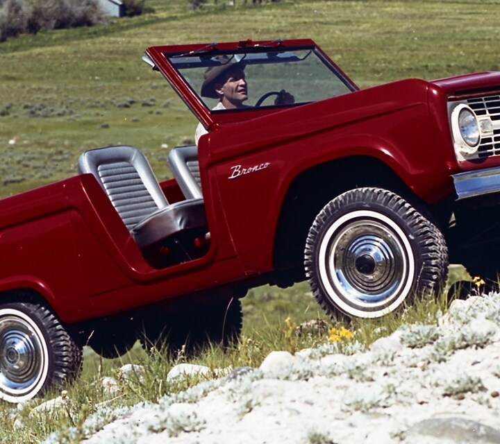 Ford Bronco Roadster 1966 en rouge Rangoon qui monte sur une pente raide près d’une haute chaîne montagneuse