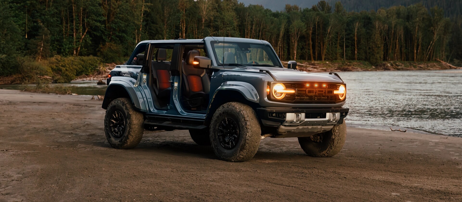 Un VUS Ford Bronco® 2025 avec les portes retirées et les lumières allumées, stationné près d’un lac dans la forêt