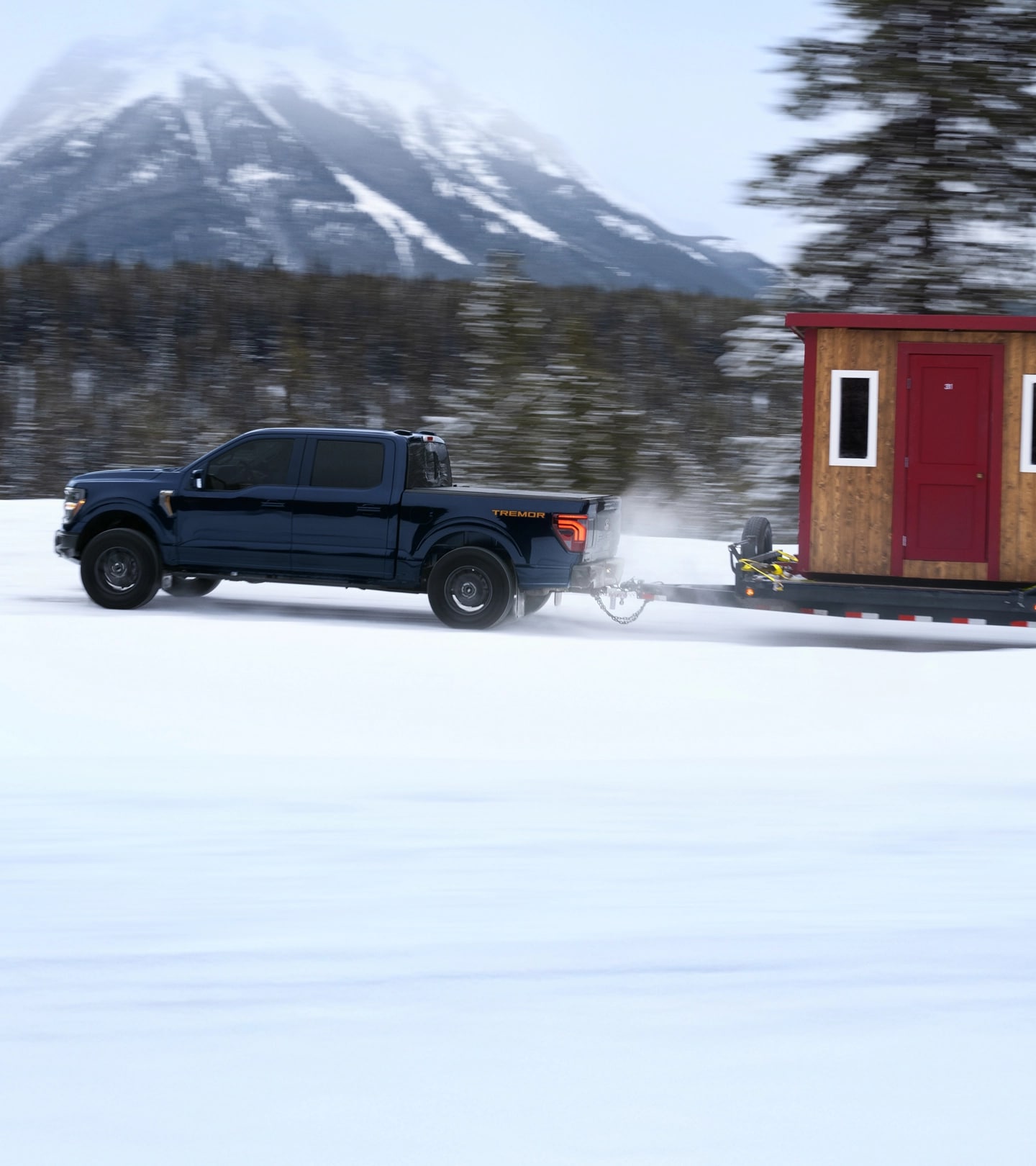 Vue de profil d’un F 150 Tremor remorquant un abri de pêche sur glace et un VTT sur une route de montagne enneigée.
