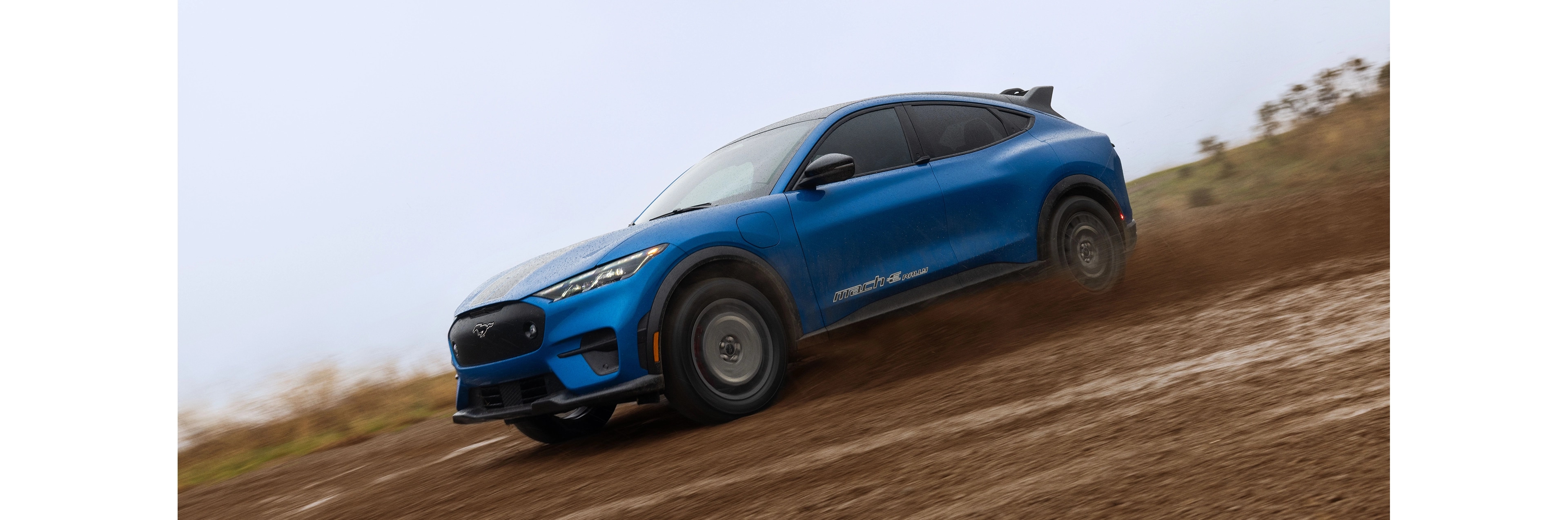 Profil latéral d'un VUS électrique Mustang Mach-E bleu roulant sur un chemin boueux.