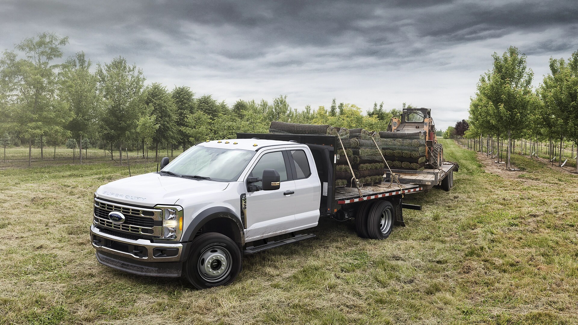 Un Ford Super Duty® à châssis-cabine blanc 2026 transportant quelque chose et tirant une remorque avec une mini-chargeuse à roues