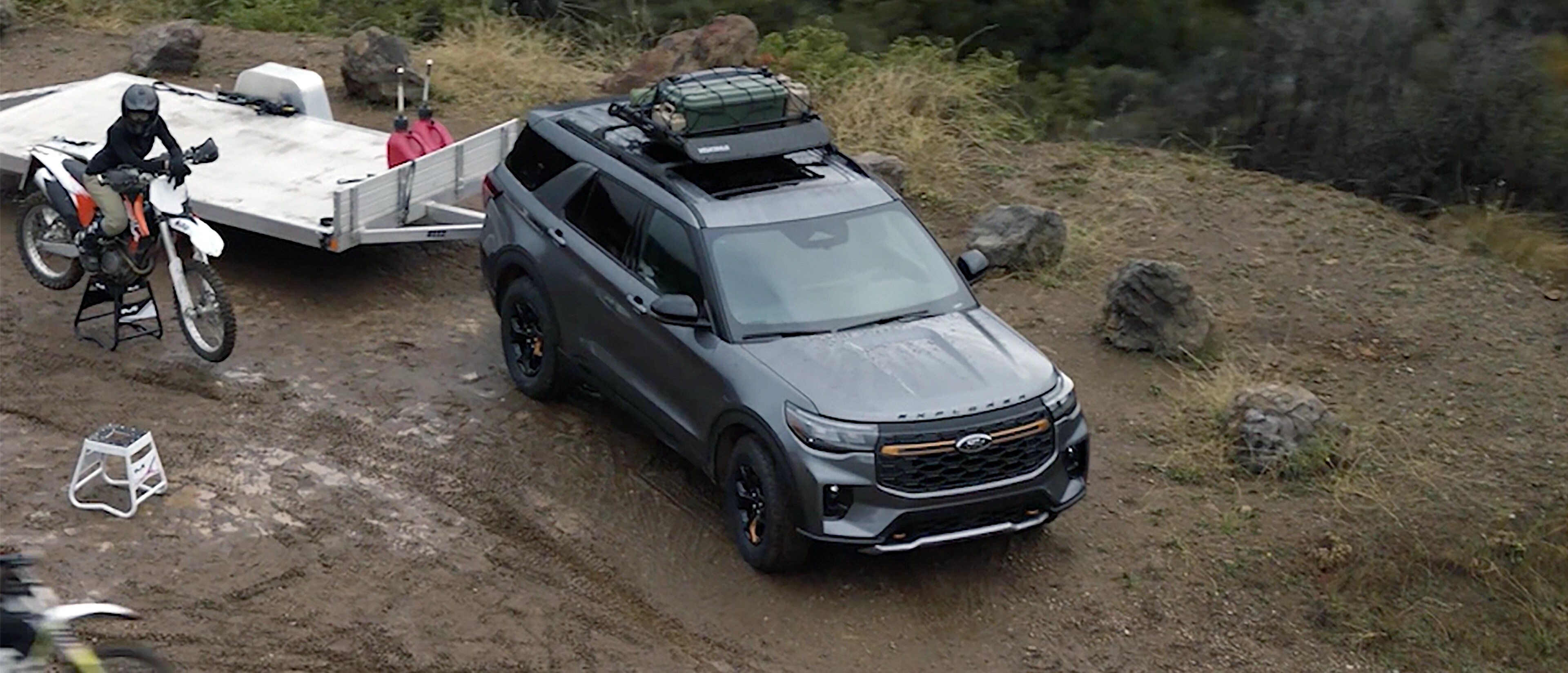 Un VUS Ford Explorer® Tremor® 2026 avec une remorque, stationnée à un point de vue montagneux, avec une personne circulant sur une moto tout-terrain à proximité