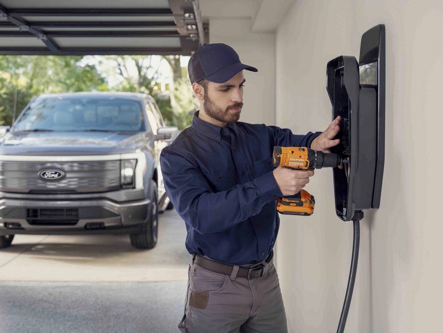 Un homme installant un poste de recharge à domicile dans un garage avec un camion Ford F-150® Lightning® 2025 stationné à l’extérieur de la porte