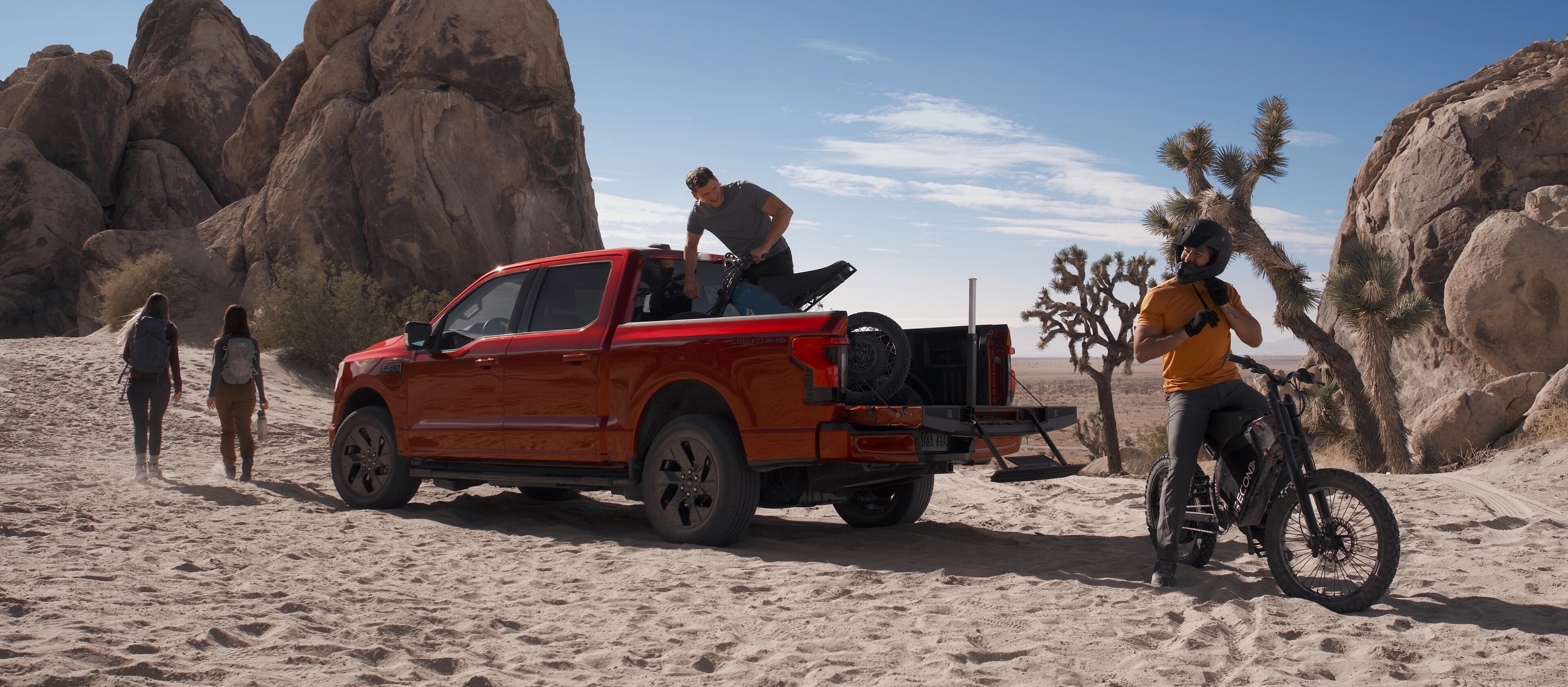 Le F-150 Lightning 2025 stationné sur du sable avec des roches et des cactus en arrière-plan. Un groupe de personnes partant en randonnée