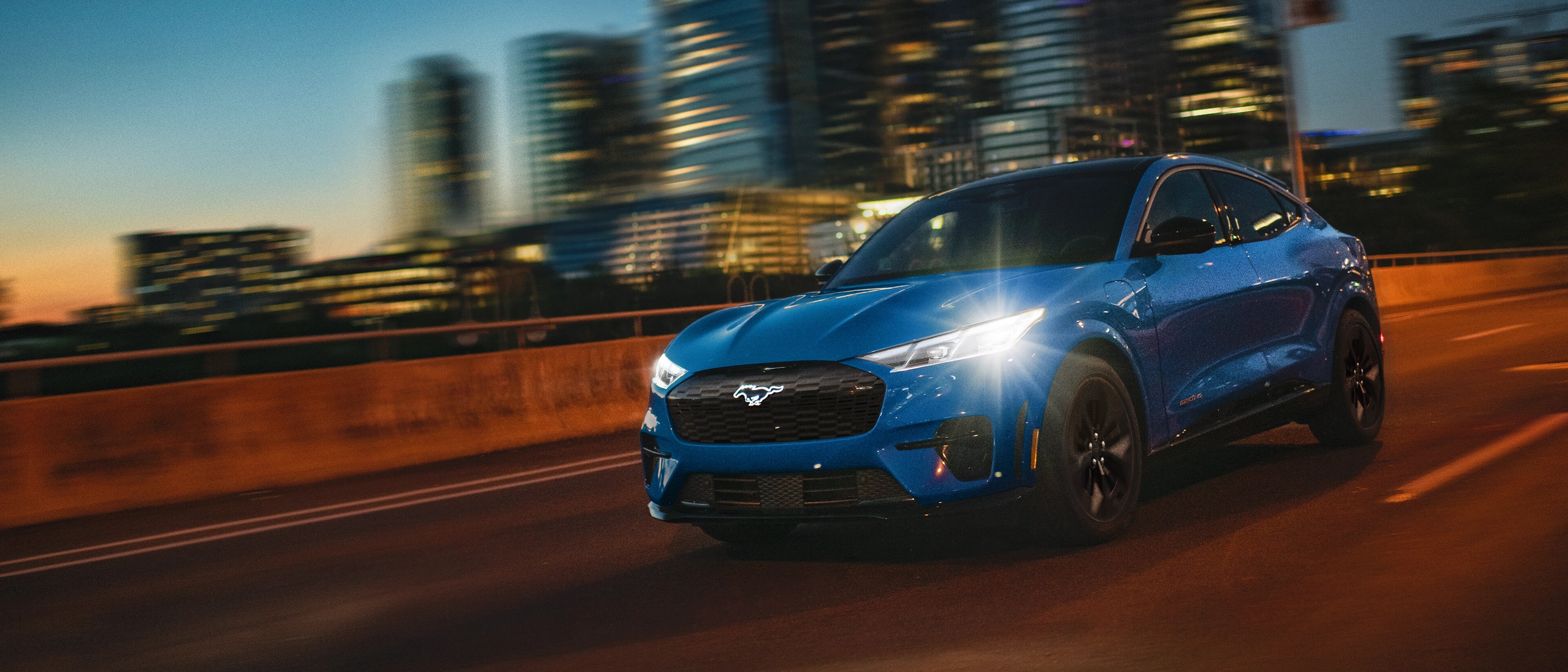 Ford Mustang Mach-E® 2025 circulant sur une autoroute la nuit avec les lumières de la ville rehaussées de reflets d’objectif