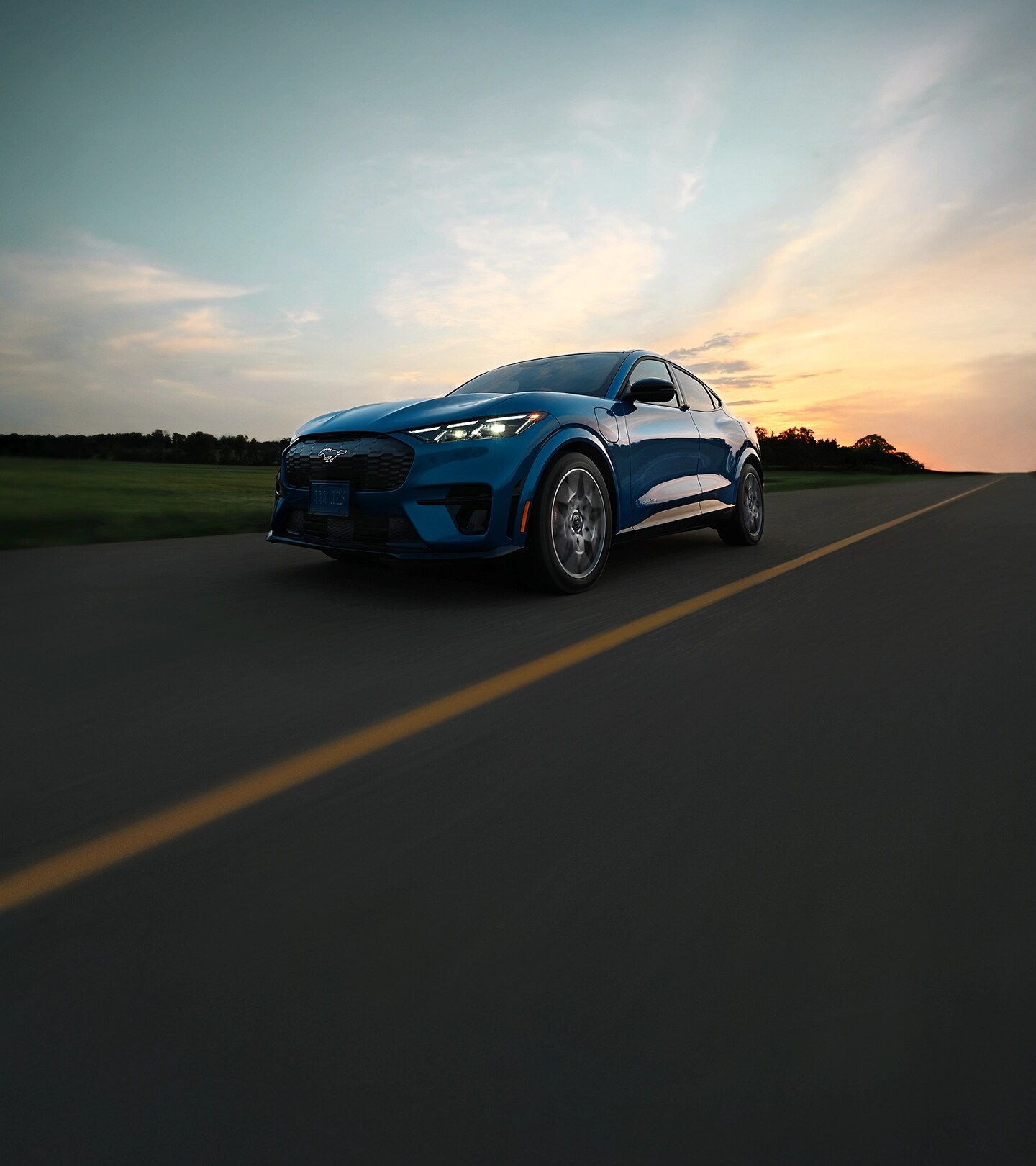 Une Ford Mustang Mach-E® GT 2025 bleue roulant sur une route ouverte au coucher du soleil, avec en arrière-plan des nuages spectaculaires et la lumière du soleil qui s'estompe