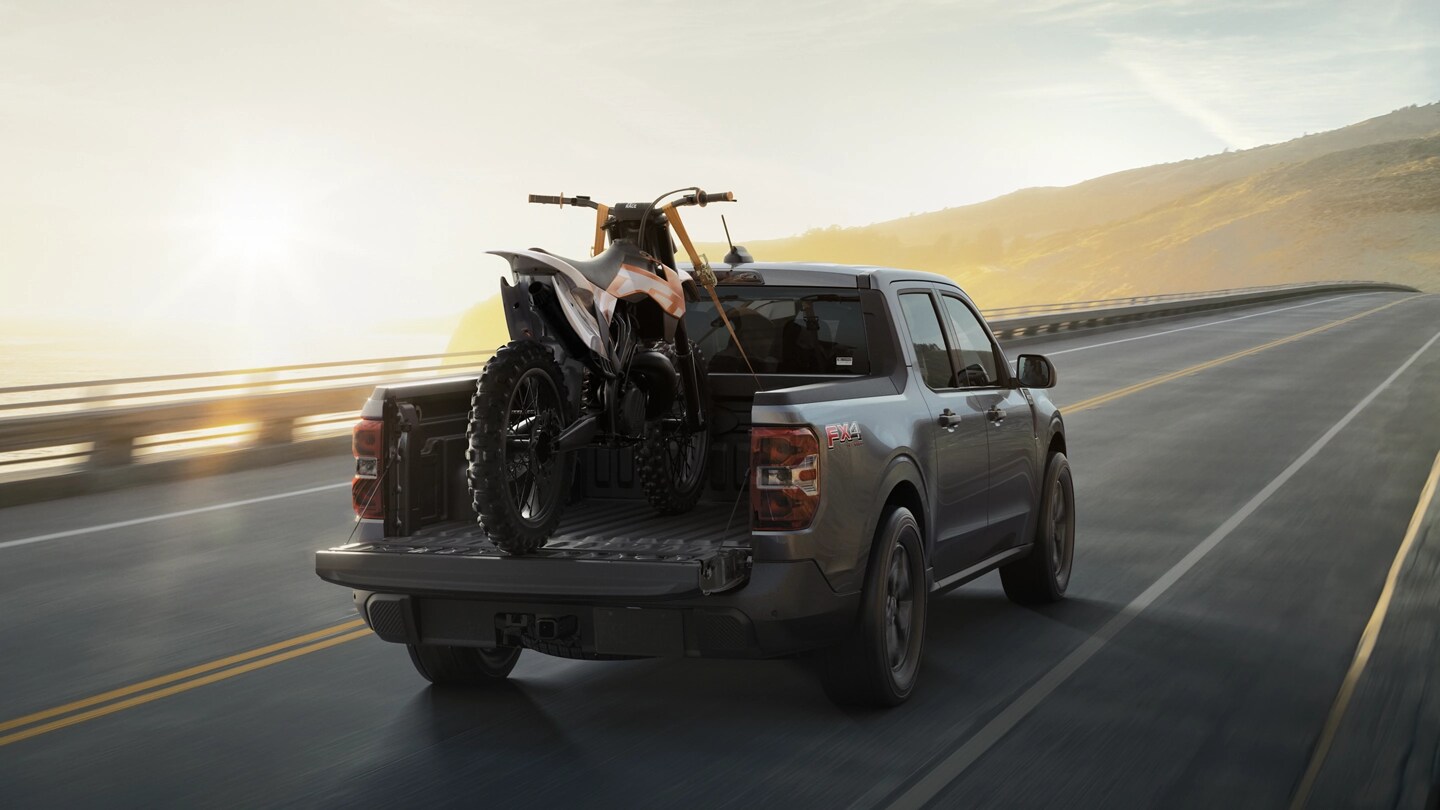 Une camionnette Ford Maverick® 2025 avec une moto tout-terrain fixée dans la caisse conduite sur une autoroute de campagne au lever du soleil