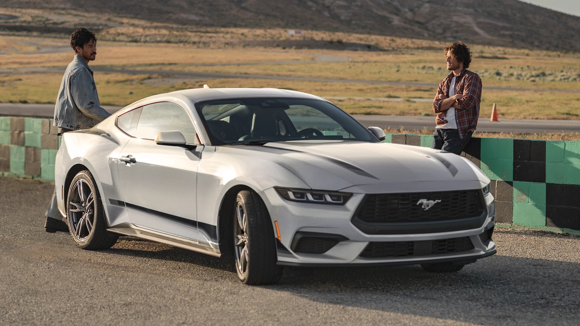 Ford Mustang® EcoBoost® Coupé 2025 avec ensemble jantes et bandes en option stationnée sur un terrain de course près de personnes