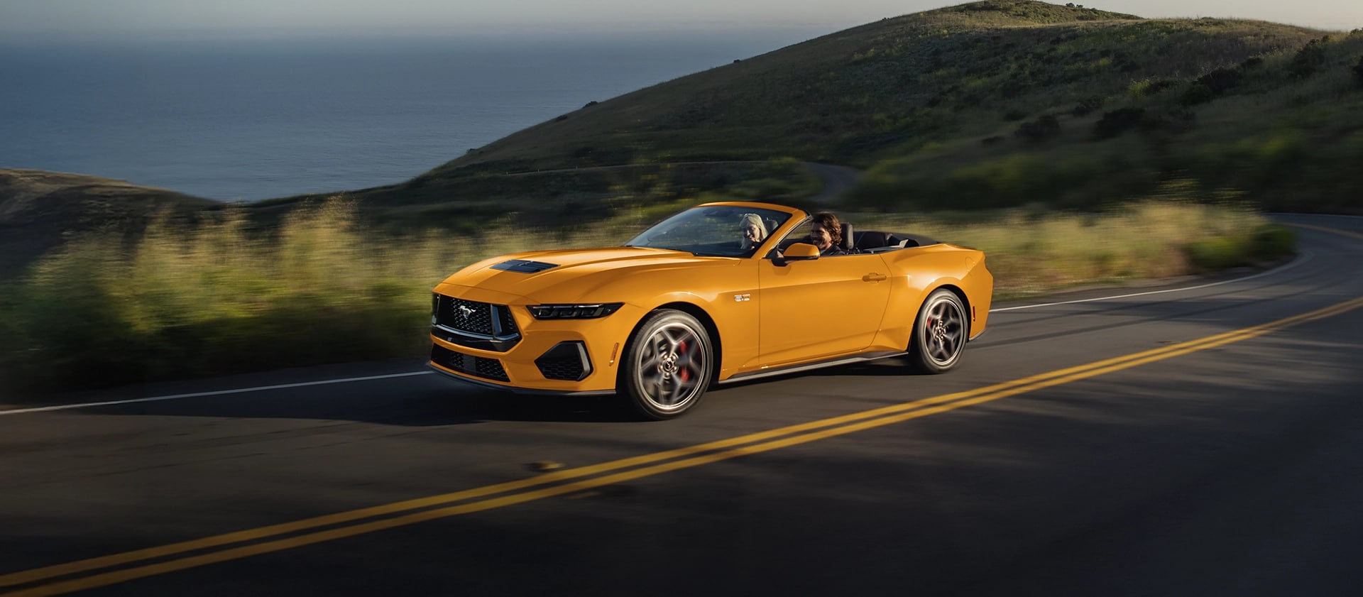 Deux personnes dans une Ford Mustang® GT décapotable 2026 couleur Furie orangée métallisée à trois couches circulant sur une autoroute côtière