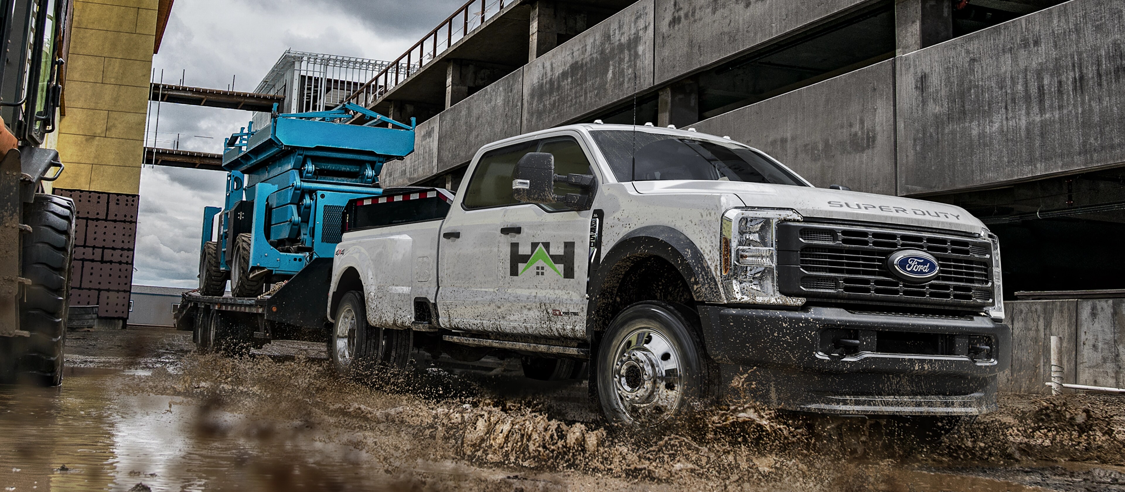 Modèle Ford Super Duty® F-450® XL 2026 tirant une remorque à plateforme avec un grand tracteur bleu dessus dans la boue et l’eau