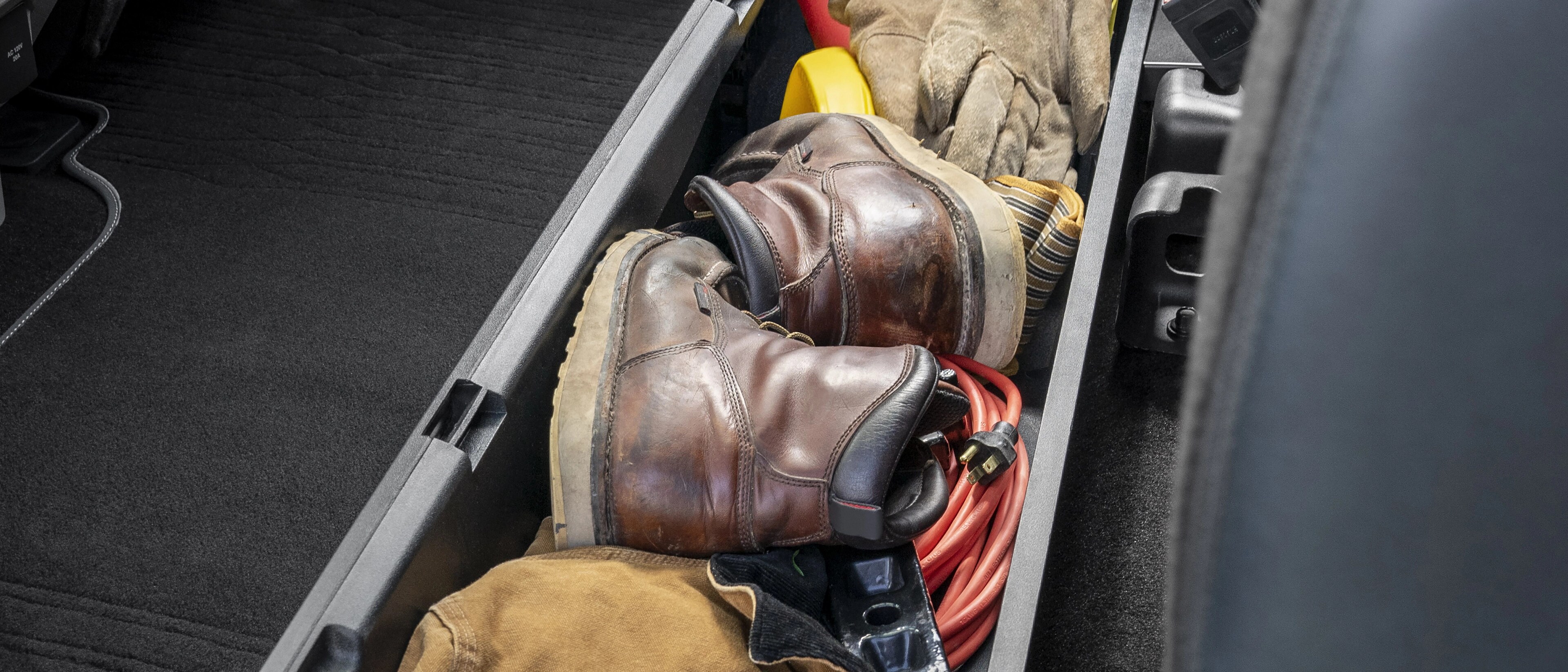 Bac de rangement sous le siège arrière retiré du siège et rempli de bottes en cuir, d’outils, de câbles et d’équipement