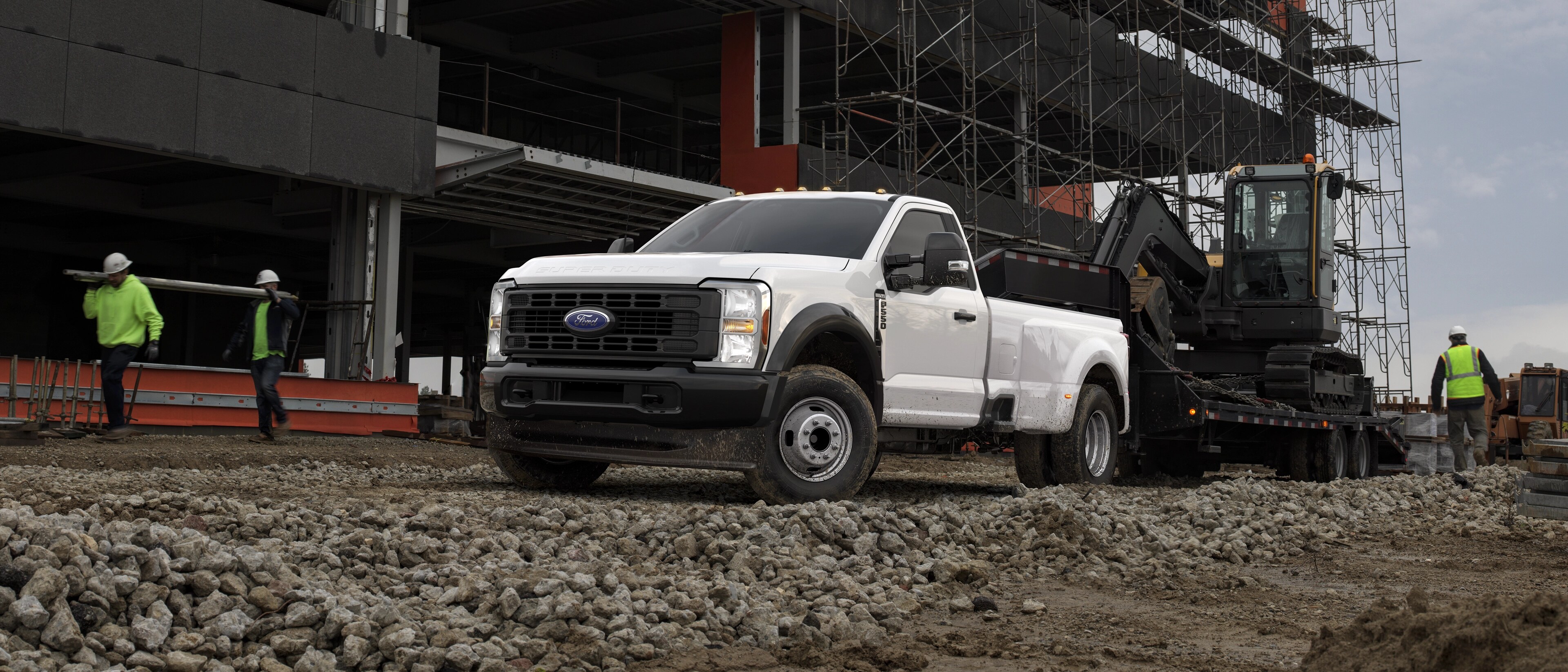 Modèle Ford Super Duty® F-350® Lariat® 2026 tirant une grande remorque à plateforme avec un tracteur lourd sur un chantier de construction