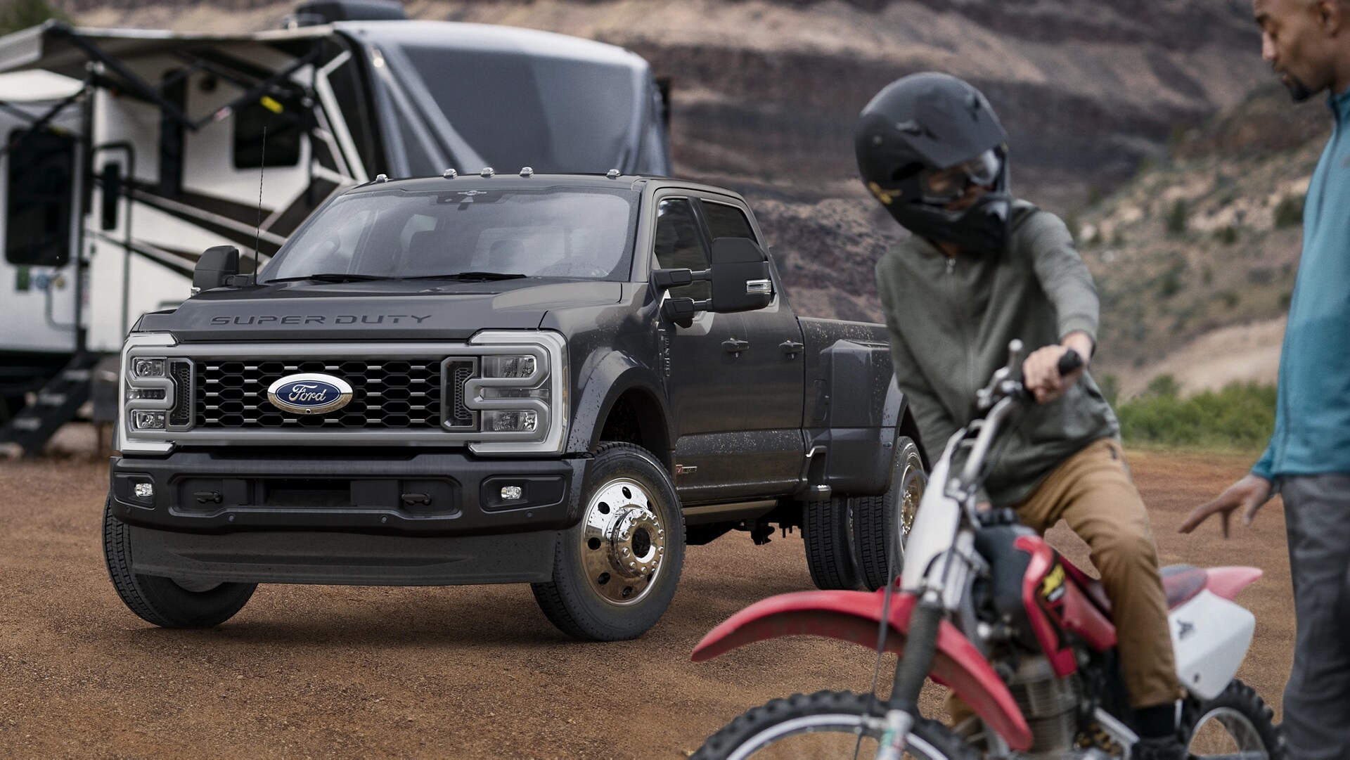 Modèle Ford Super Duty® F-450® Platinum® 2026 en gris carbonisé stationné sur un terrain de camping avec des caravanes en arrière-plan et une personne sur un vélo en avant-plan.