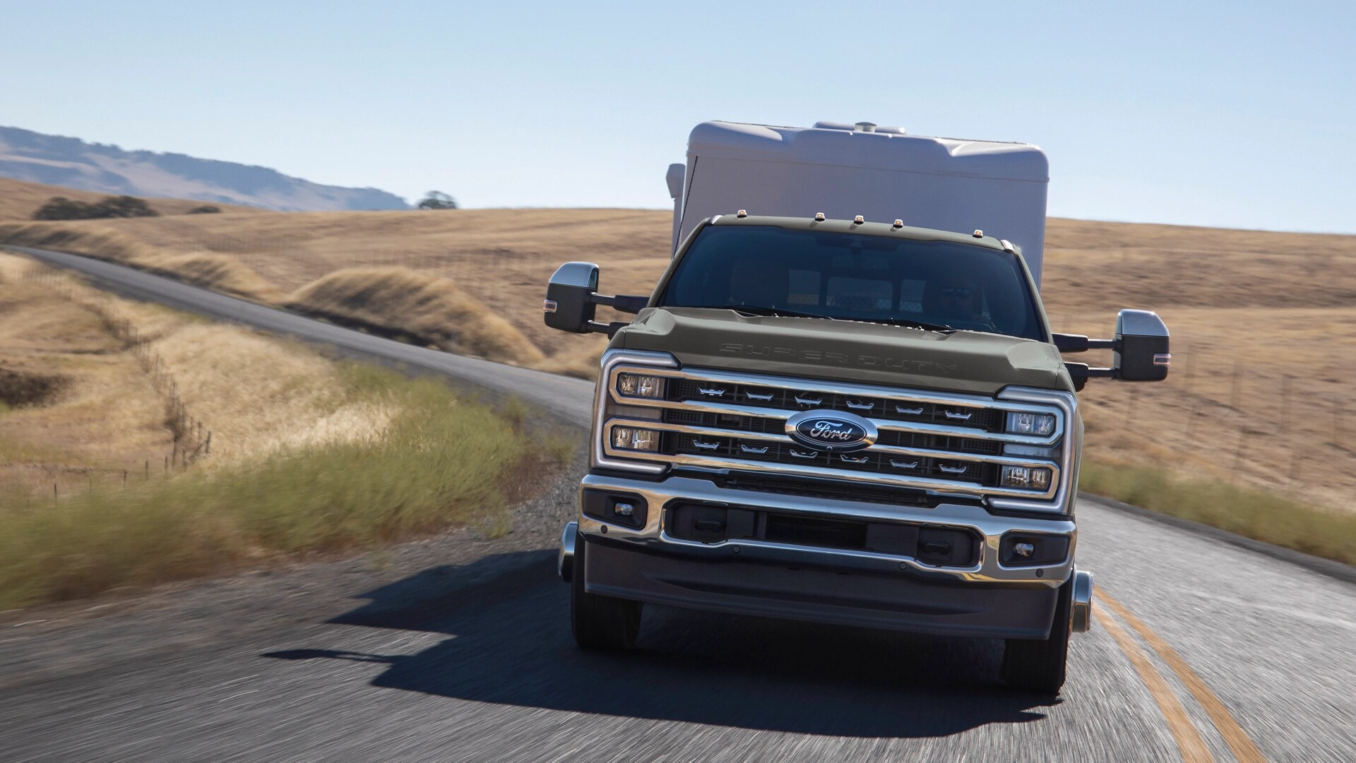Modèle Ford Super Duty® F-350® Lariat® 2026 avec ensemble chromé circulant sur une route pavée vallonnée tout en tirant une grande remorque