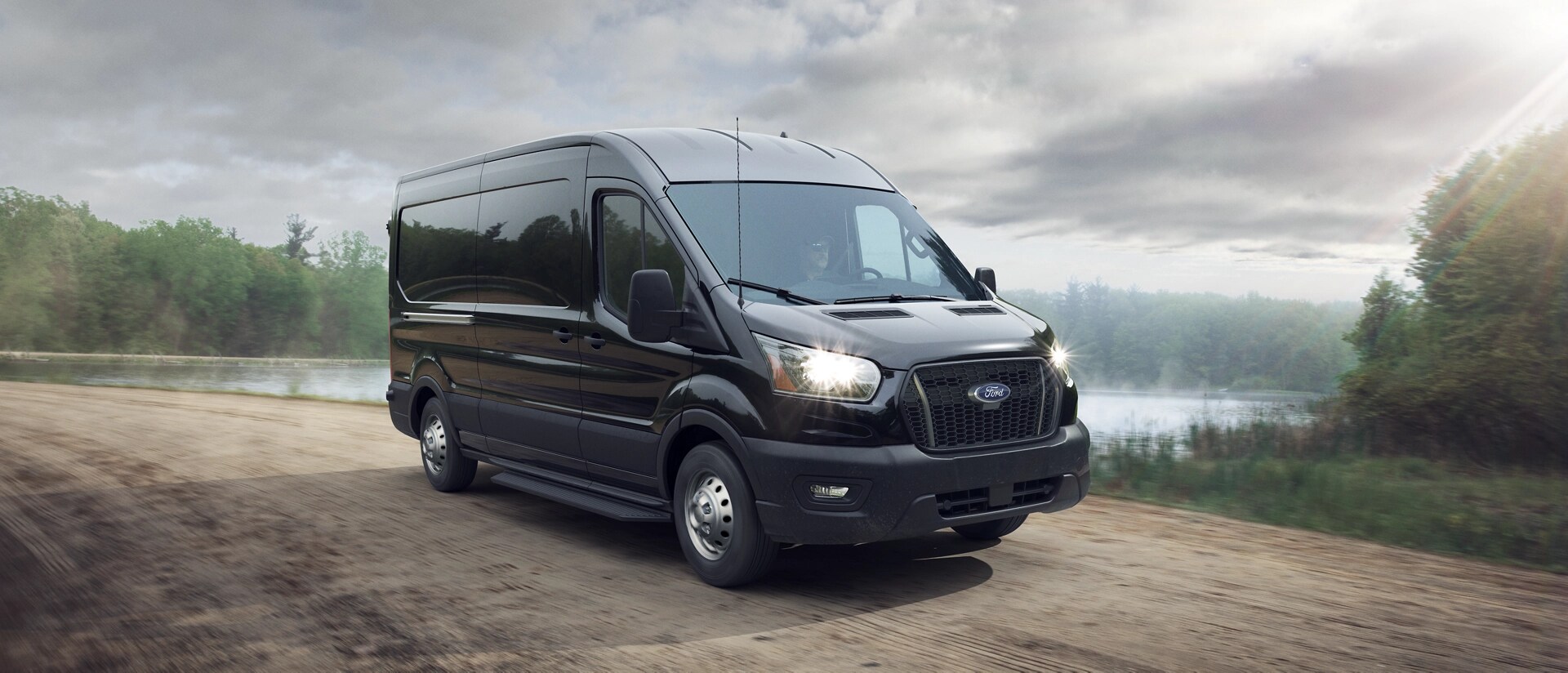 Une fourgonnette Ford Transit® 2025 conduite sur un chemin de terre près d’un plan d’eau