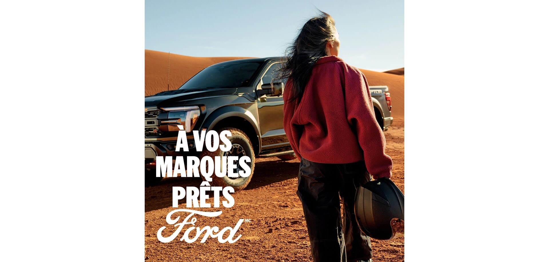 Une femme, dos tourné, tient un casque alors qu'elle est debout dans le désert devant un Ford F-150 Raptor gris foncé. Le mot « Ford » en blanc dans le coin gauche. Le texte blanc « À vos marques. Prêts.Ford » dans le coin inférieur droit