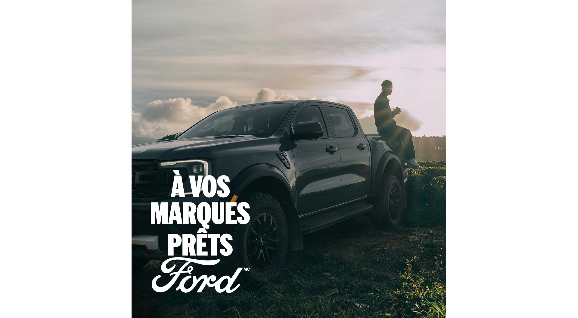 Vue de trois quarts d'un Ford Ranger Raptor gris garé sur l'herbe. Une personne est assise à l'arrière du pick-up, le soleil brille en arrière-plan. À vos marques, prêts, Ford.