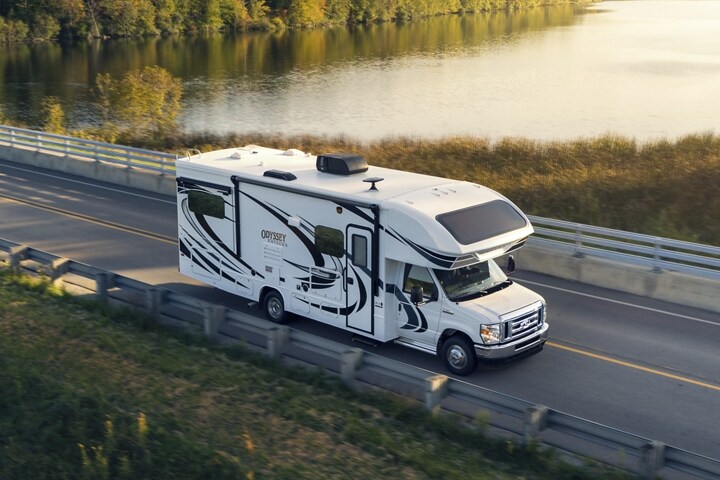 Fourgon tronqué à roues arrière jumelées Ford série E 2025 avec autocaravane de classe C circulant près d’un grand plan d’eau
