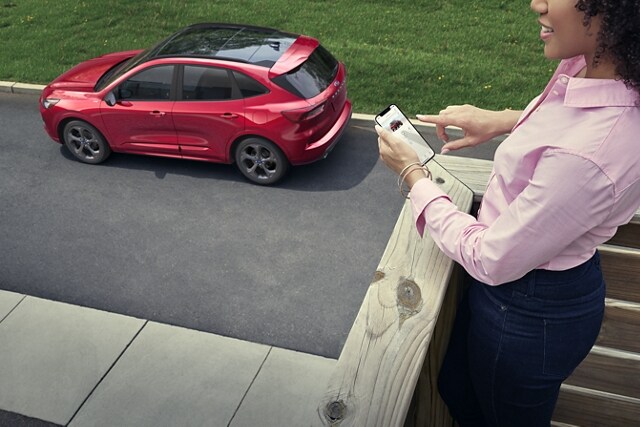 La personne tient un téléphone intelligent sur un porche surplombant un Ford Escape® 2023 en rouge rapide teint métallisé (en sus)
