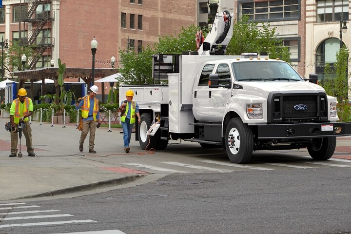 Travailleurs marchant près d’un Ford F-750 2025 à cabine six places avec aménagement de nacelle élévatrice, couleur blanc Oxford et stationné dans une rue de la ville