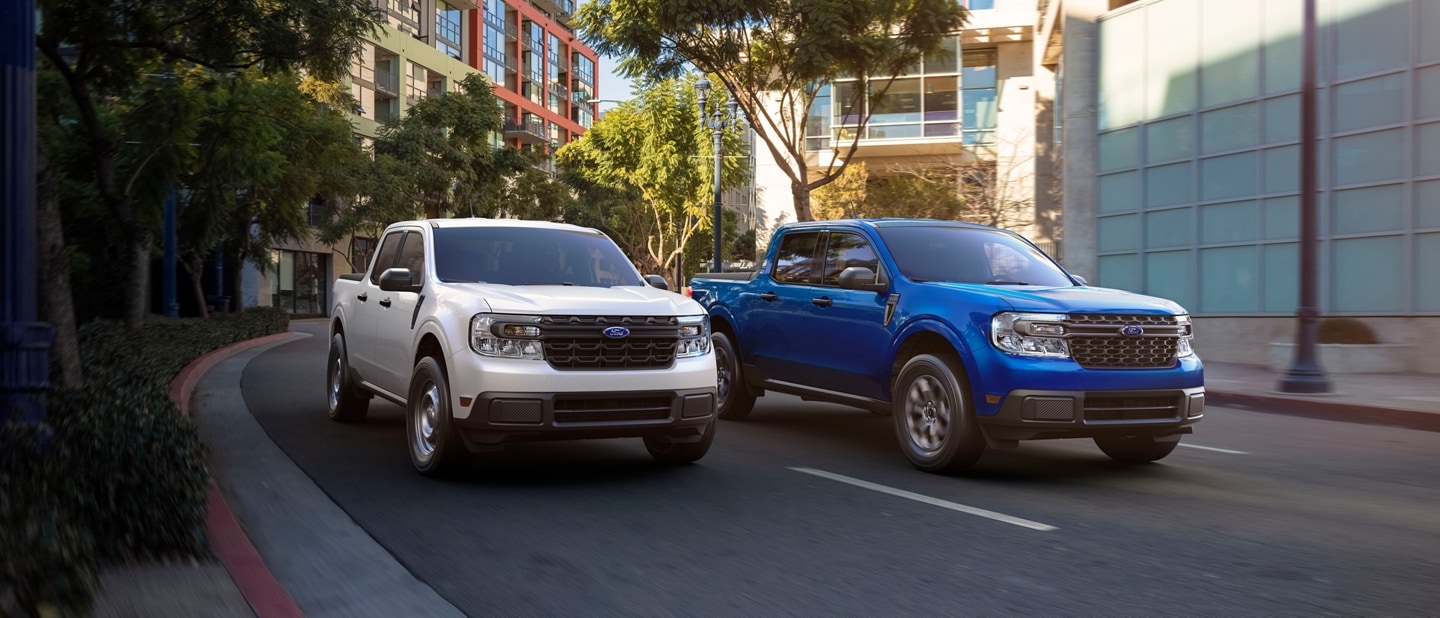 Deux camions Ford Maverick® 2024 en blanc Oxford et bleu atlas métallisé sont conduits sur une route urbaine bordée d’arbres
