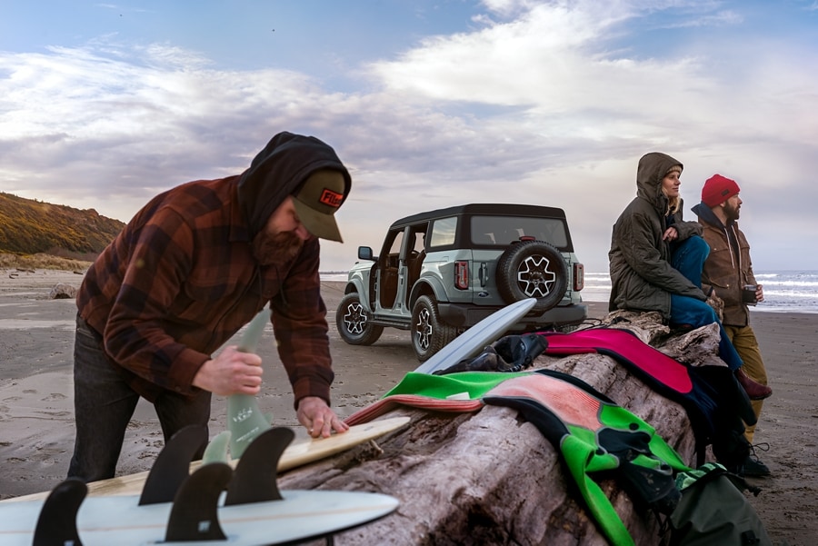 Modèle Ford Bronco® Outer Banks™ 2023 en gris cactus sur la plage avec trois surfeurs